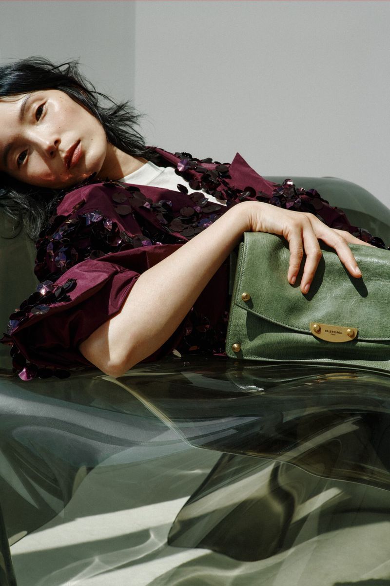 Model Xiao Wen Ju reclining in a sequined burgundy jacket holding a green leather clutch bag.