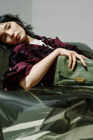 Model Xiao Wen Ju reclining in a sequined burgundy jacket holding a green leather clutch bag.