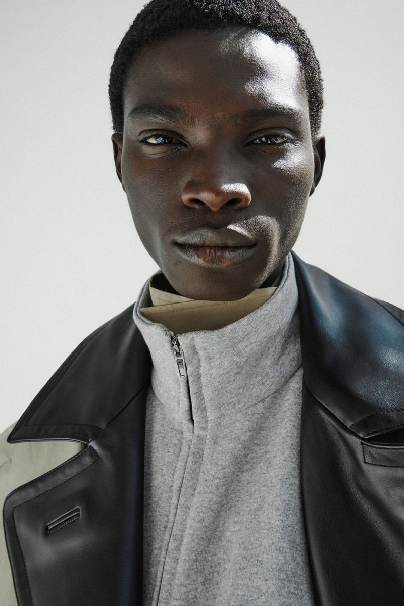 Close-up portrait of a Black male model wearing a grey zip-up jacket and black leather collar against an off-white background.