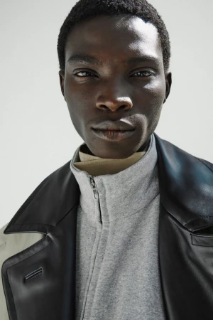 Close-up portrait of a Black male model wearing a grey zip-up jacket and black leather collar against an off-white background.