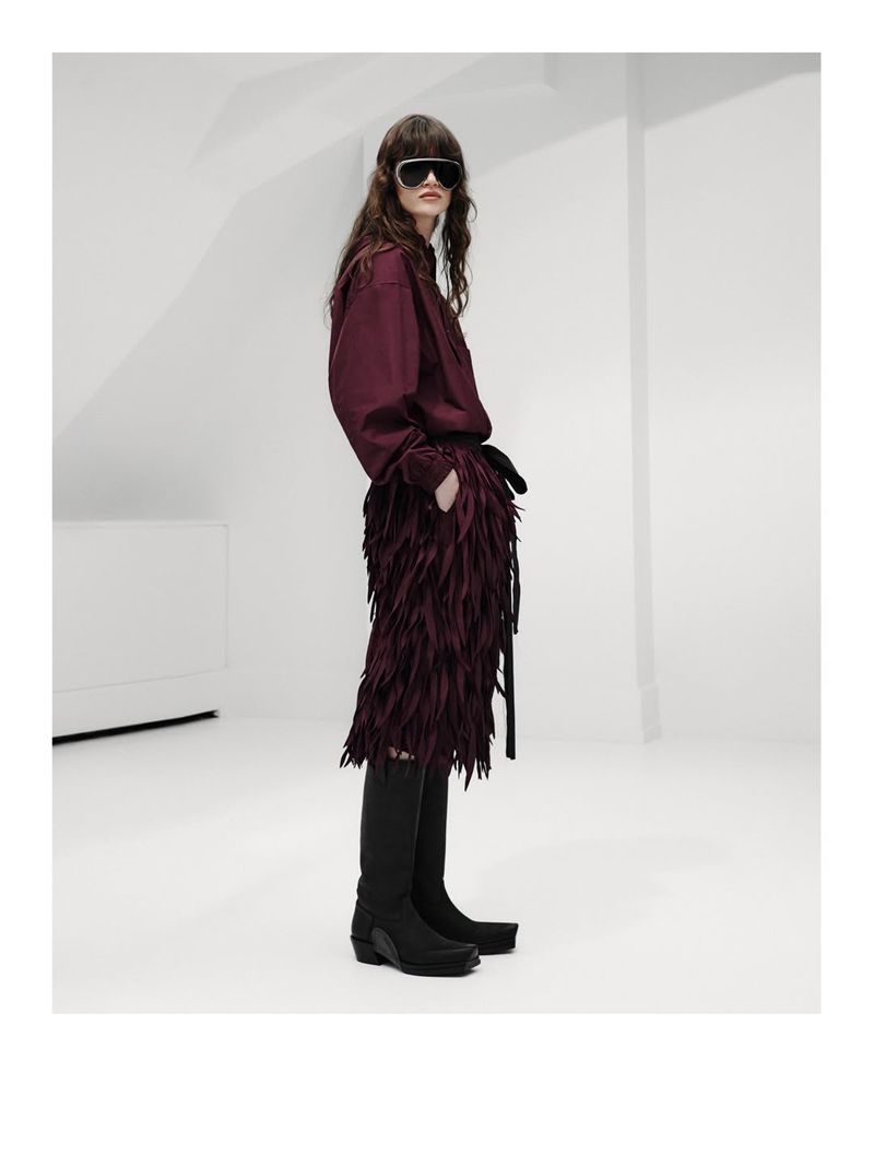 Model wearing a burgundy shirt, matching fringe skirt, black shield sunglasses, and tall black boots against a white studio setting.