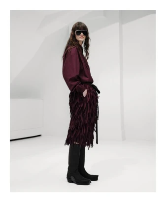 Model wearing a burgundy shirt, matching fringe skirt, black shield sunglasses, and tall black boots against a white studio setting.