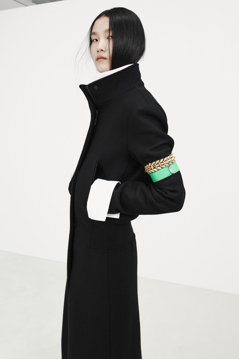 Model wearing a tailored black coat with an green armband and chunky gold chain accessory against a white background.