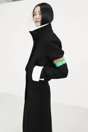 Model wearing a tailored black coat with an green armband and chunky gold chain accessory against a white background.