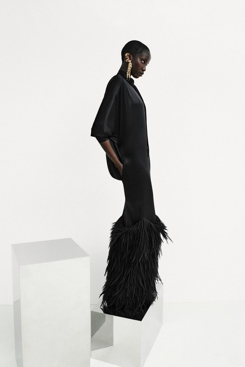 Model Anok Yai in a black satin feather-hem dress with a gold ear cuff, posing beside mirrored pedestals against a white backdrop.