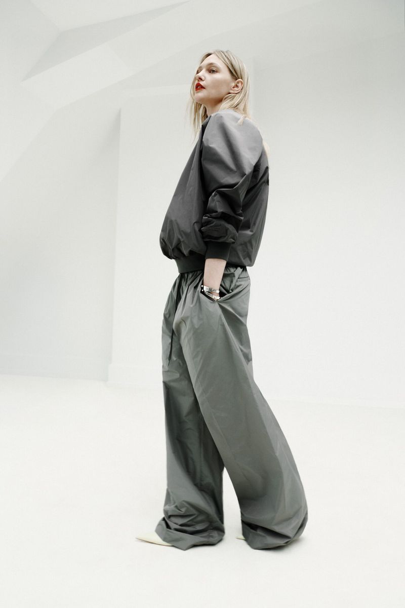 Model in voluminous dark grey bomber jacket and matching wide-leg trousers posing against a clean white architectural background.
