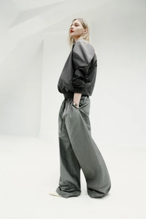Model in voluminous dark grey bomber jacket and matching wide-leg trousers posing against a clean white architectural background.