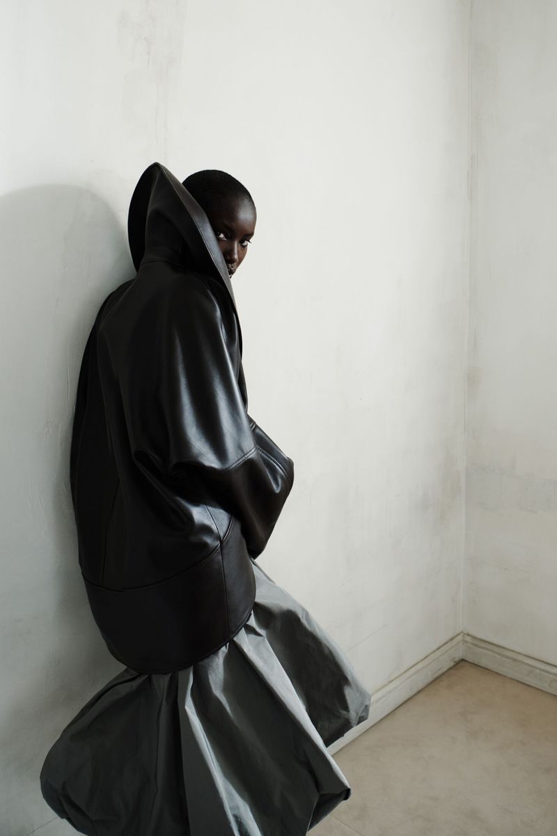 Anok Yai wearing an oversized black sculptural hooded leather jacket and grey balloon skirt against a white wall.