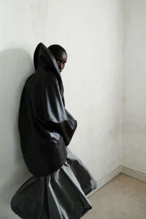 Anok Yai wearing an oversized black sculptural hooded leather jacket and grey balloon skirt against a white wall.