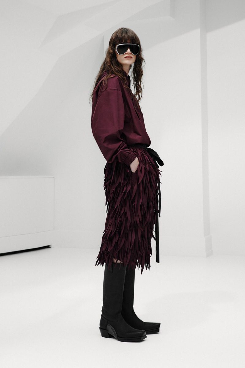 Model wearing dark visor sunglasses, a burgundy shirt, textured fringe skirt, and black knee-high boots against a white studio set.