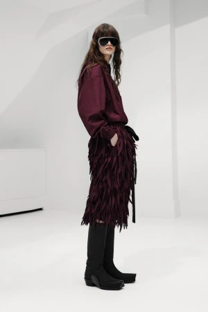 Model wearing dark visor sunglasses, a burgundy shirt, textured fringe skirt, and black knee-high boots against a white studio set.