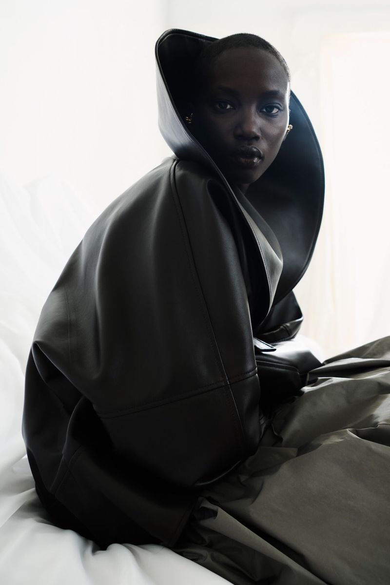 Model Anok Yai wearing an oversized sculptural black leather coat with a dramatic high collar against a white backdrop.