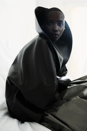 Model Anok Yai wearing an oversized sculptural black leather coat with a dramatic high collar against a white backdrop.