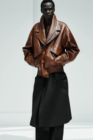 Model Malick Bodian wearing an oversized brown leather jacket and layered black coat against a white studio background.