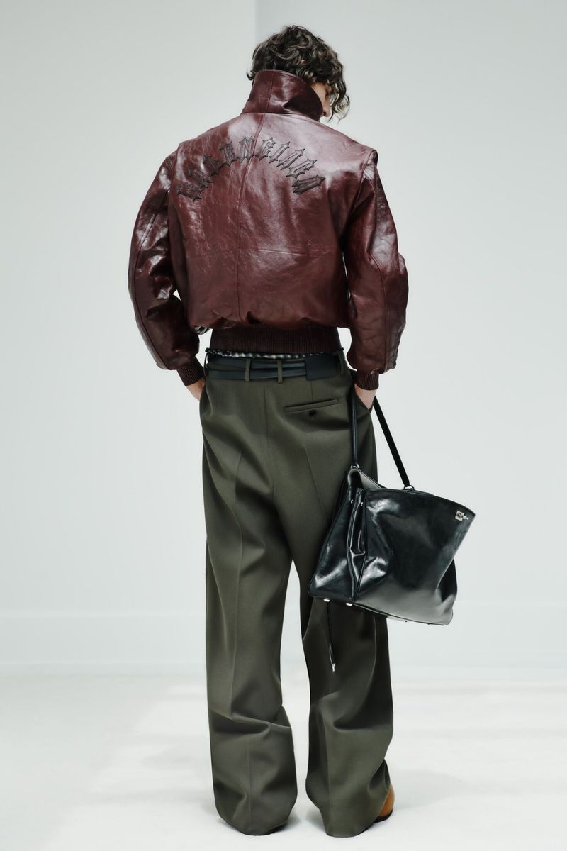 Model seen from behind wearing a burgundy leather bomber jacket, olive wide-leg trousers, and holding a glossy black leather bag.