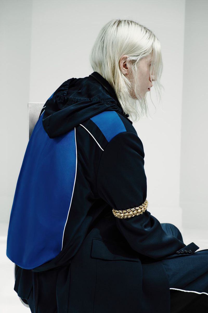 Model with platinum blonde hair wearing a black and blue hooded jacket with a gold chain armband against a white backdrop.