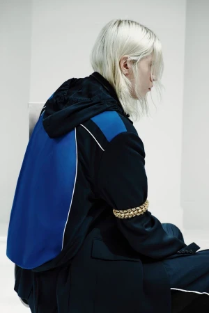Model with platinum blonde hair wearing a black and blue hooded jacket with a gold chain armband against a white backdrop.