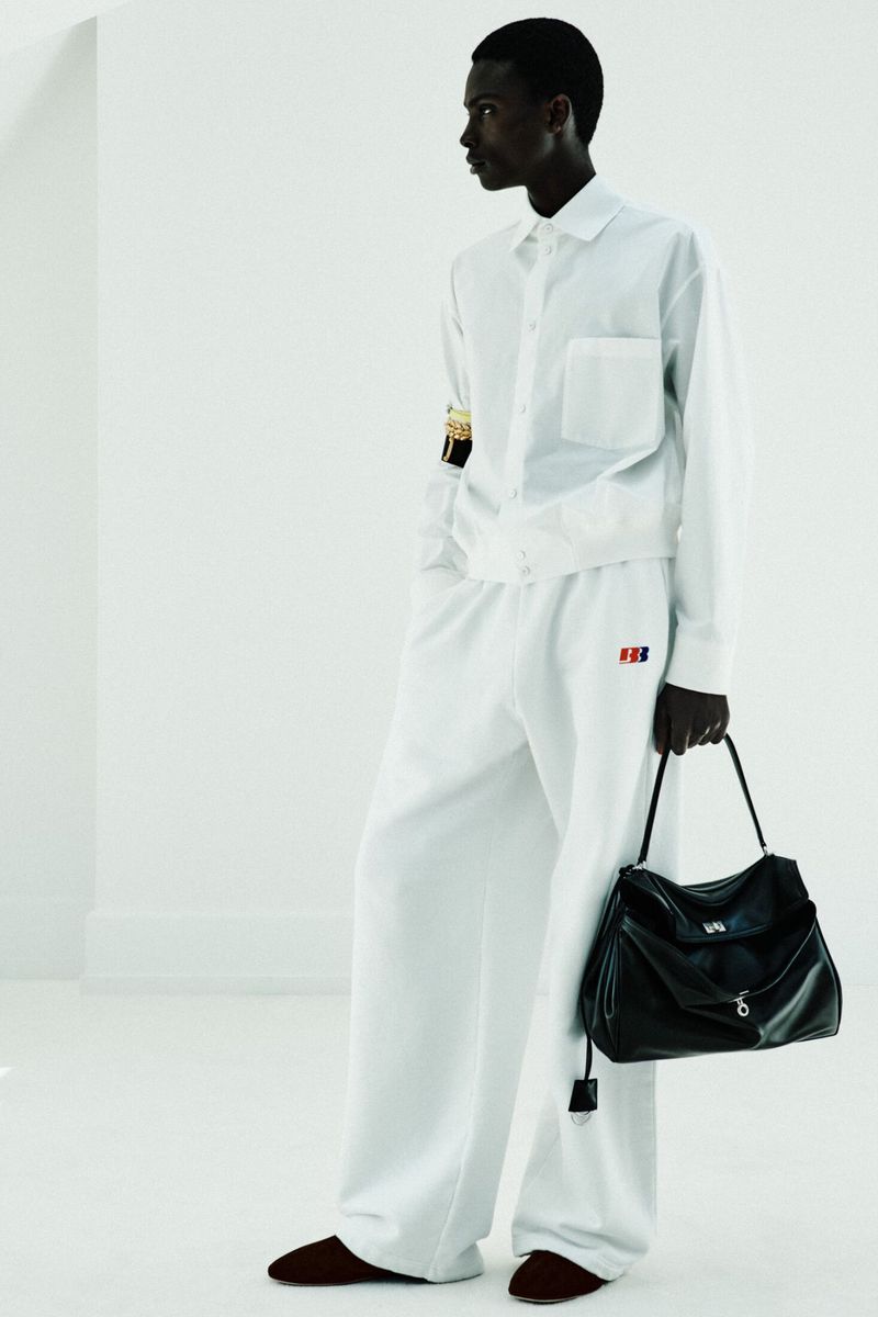 Model wearing an all-white button-up shirt and wide-leg trousers holding a black leather bag against a bright white background.