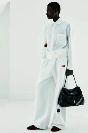 Model wearing an all-white button-up shirt and wide-leg trousers holding a black leather bag against a bright white background.