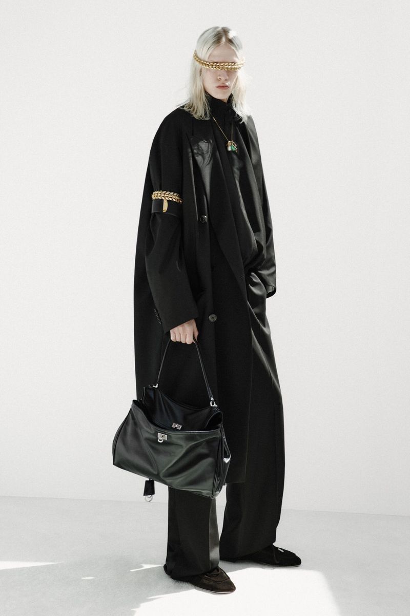 Model wearing an oversized black coat and trousers holding a black leather bag against a clean white background.