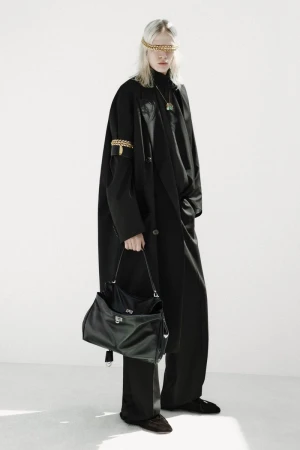 Model wearing an oversized black coat and trousers holding a black leather bag against a clean white background.
