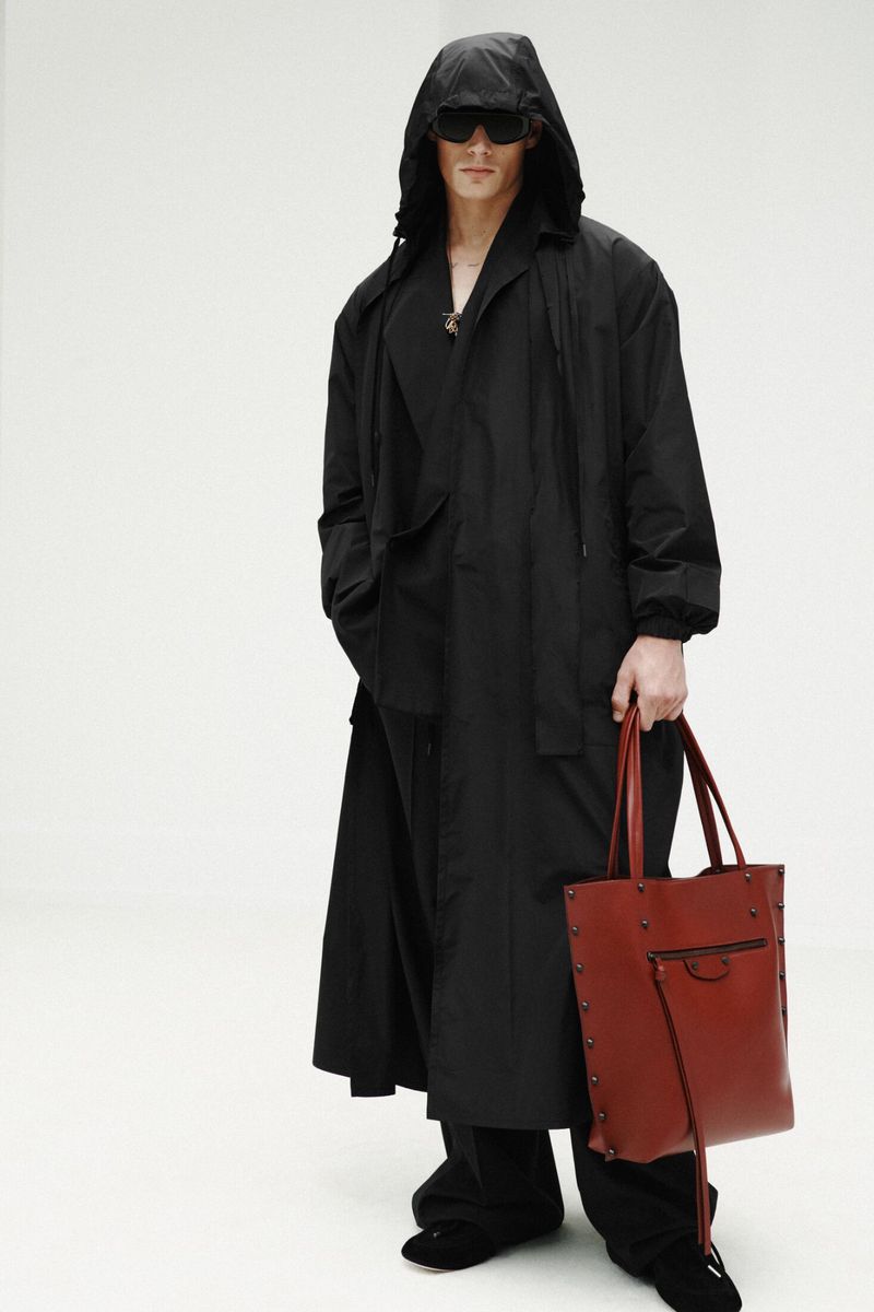 Model wearing an oversized hooded black coat and dark sunglasses holding a reddish-brown leather tote bag against a white backdrop.