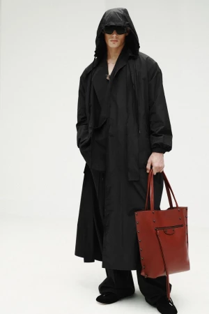 Model wearing an oversized hooded black coat and dark sunglasses holding a reddish-brown leather tote bag against a white backdrop.