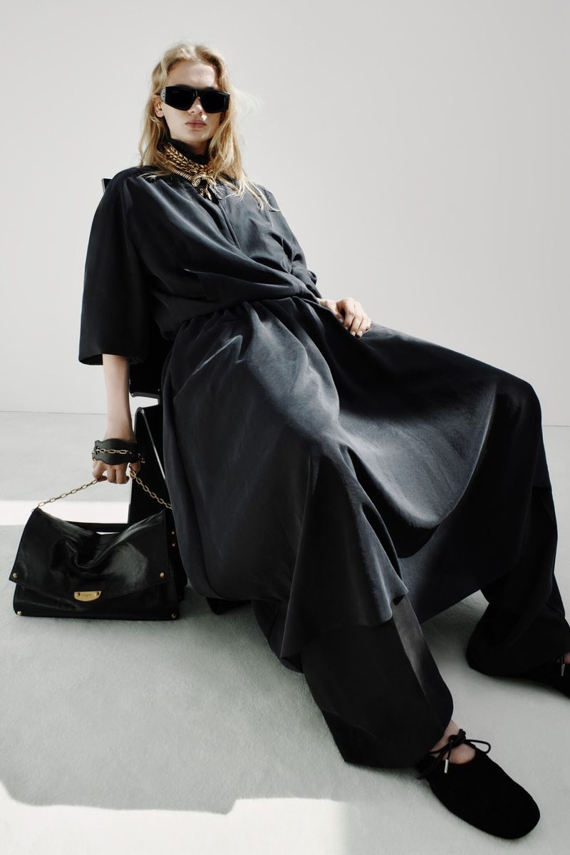 Model Sasha Pivovarova seated in an oversized black draped outfit with sunglasses and a black leather handbag in a white studio.