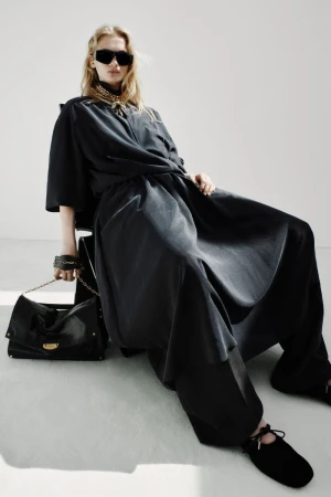 Model Sasha Pivovarova seated in an oversized black draped outfit with sunglasses and a black leather handbag in a white studio.