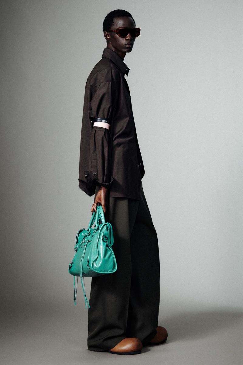 Model wearing dark oversized shirt and trousers holding a vibrant turquoise handbag against a grey studio backdrop.