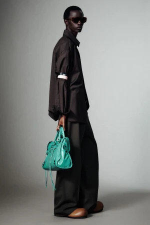 Model wearing dark oversized shirt and trousers holding a vibrant turquoise handbag against a grey studio backdrop.
