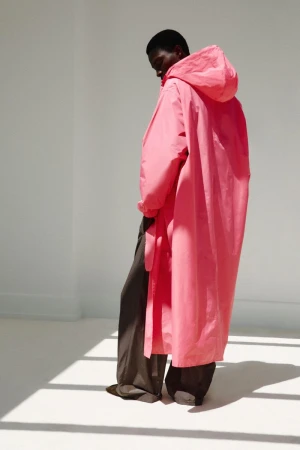 Model wearing an oversized pink hooded raincoat and dark wide-leg trousers in a minimalist white studio.