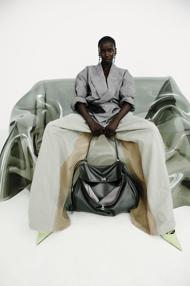 Anok Yai seated on a translucent sculptural chair holding a black leather handbag in a grey tailored jacket and wide-leg trousers.