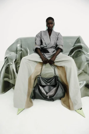 Anok Yai seated on a translucent sculptural chair holding a black leather handbag in a grey tailored jacket and wide-leg trousers.