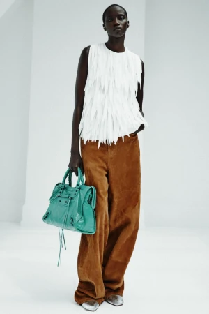 Anok Yai wears a textured white sleeveless top, brown wide-leg pants, and carries a turquoise handbag against a white studio background.
