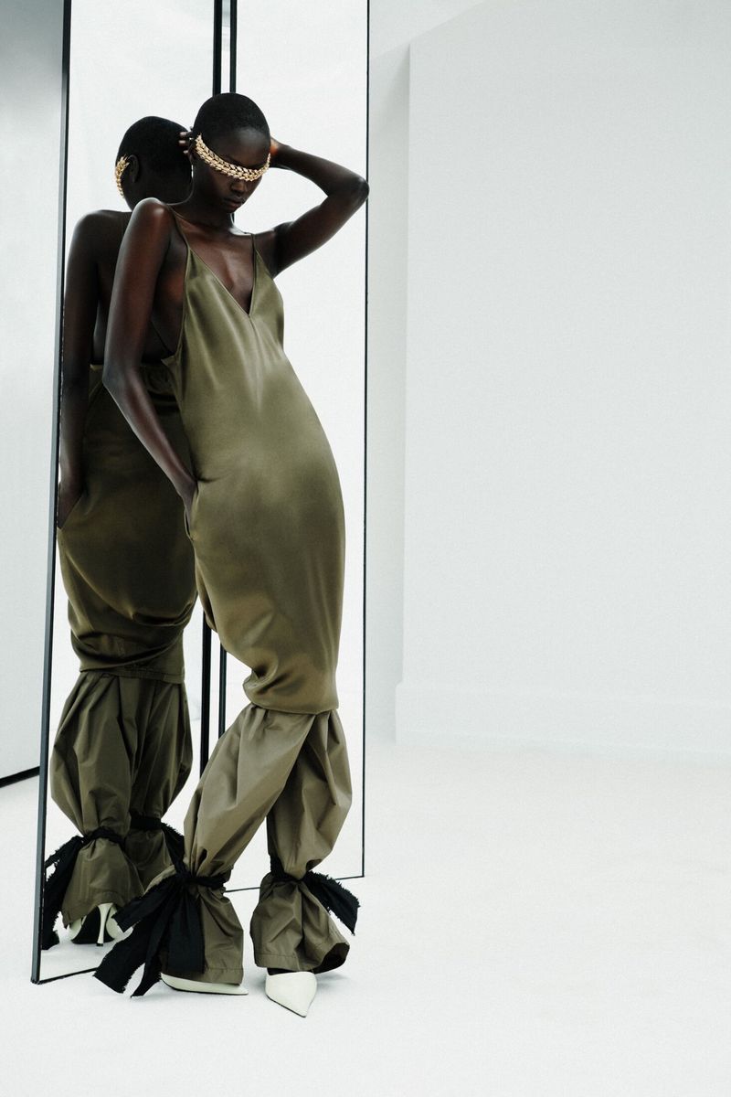 Model wearing an olive green slip dress with tied details and face jewelry standing against a white backdrop.