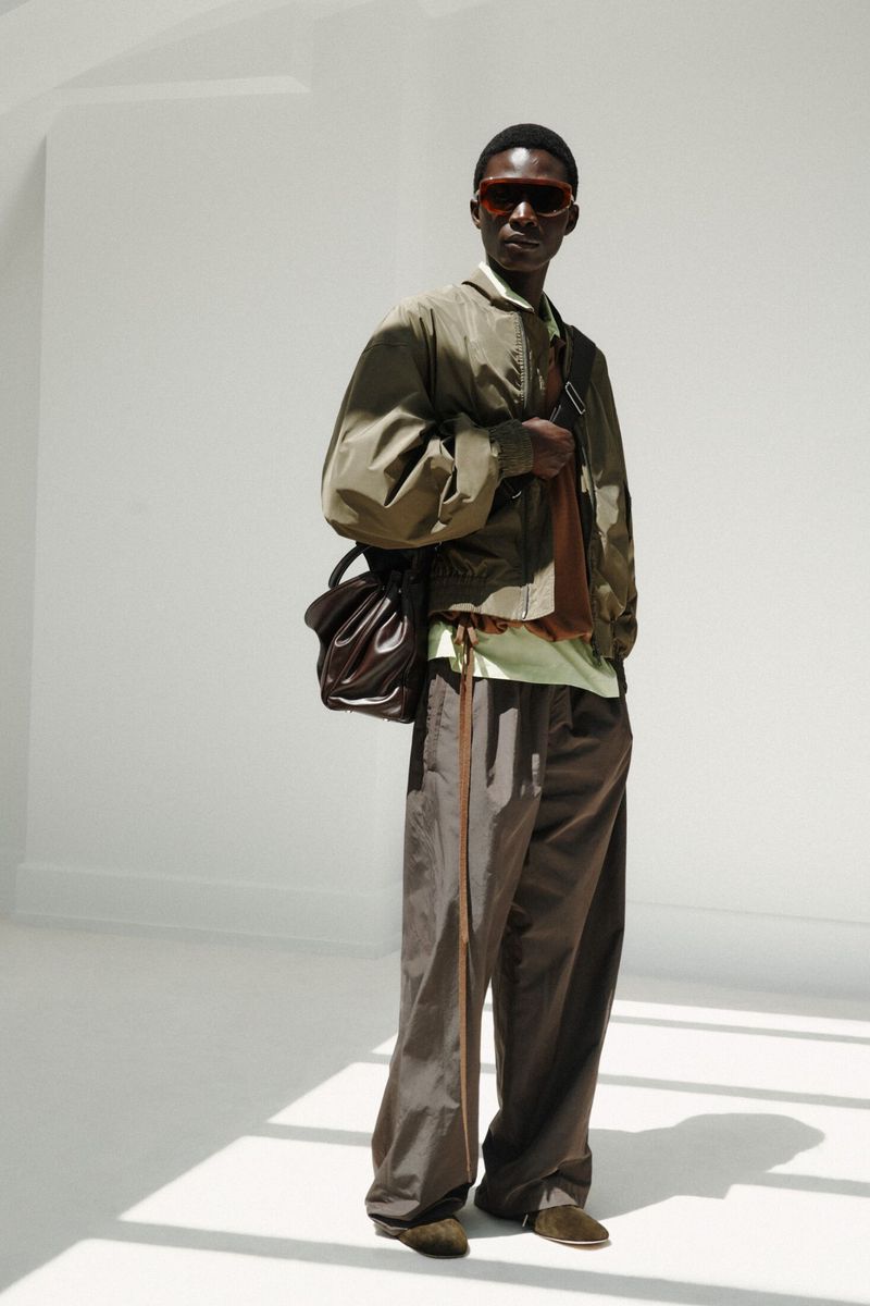 Model wearing dark sunglasses, an olive bomber jacket, and brown trousers against a bright white studio backdrop