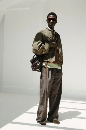 Model wearing dark sunglasses, an olive bomber jacket, and brown trousers against a bright white studio backdrop