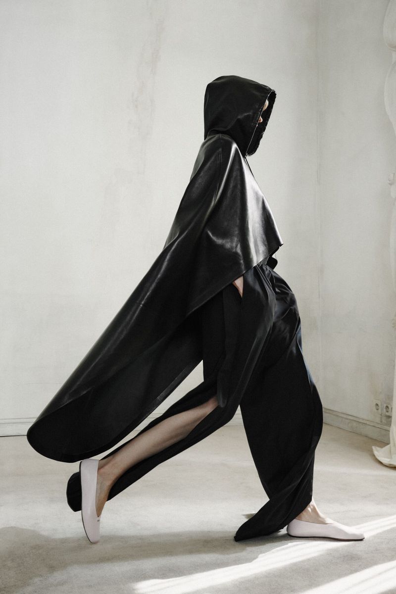 Model walking in profile wearing a black hooded leather cape, loose black trousers, and pale flats against a light interior wall.