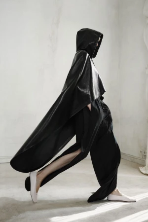 Model walking in profile wearing a black hooded leather cape, loose black trousers, and pale flats against a light interior wall.