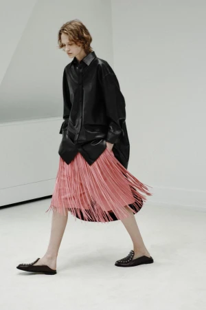 Model walking in a black leather shirt and pink fringed skirt against a white studio background.