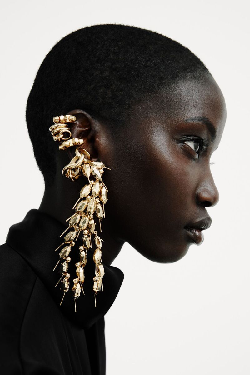 Profile of Anok Yai with short buzzed hair wearing elaborate gold drop ear jewelry and a black top against a white backdrop.
