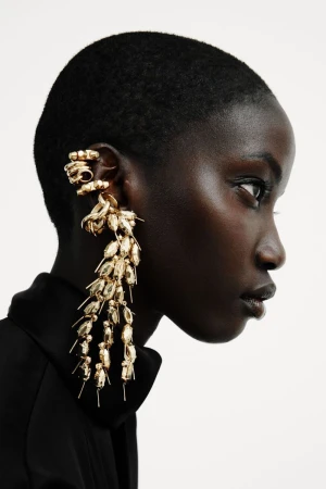 Profile of Anok Yai with short buzzed hair wearing elaborate gold drop ear jewelry and a black top against a white backdrop.