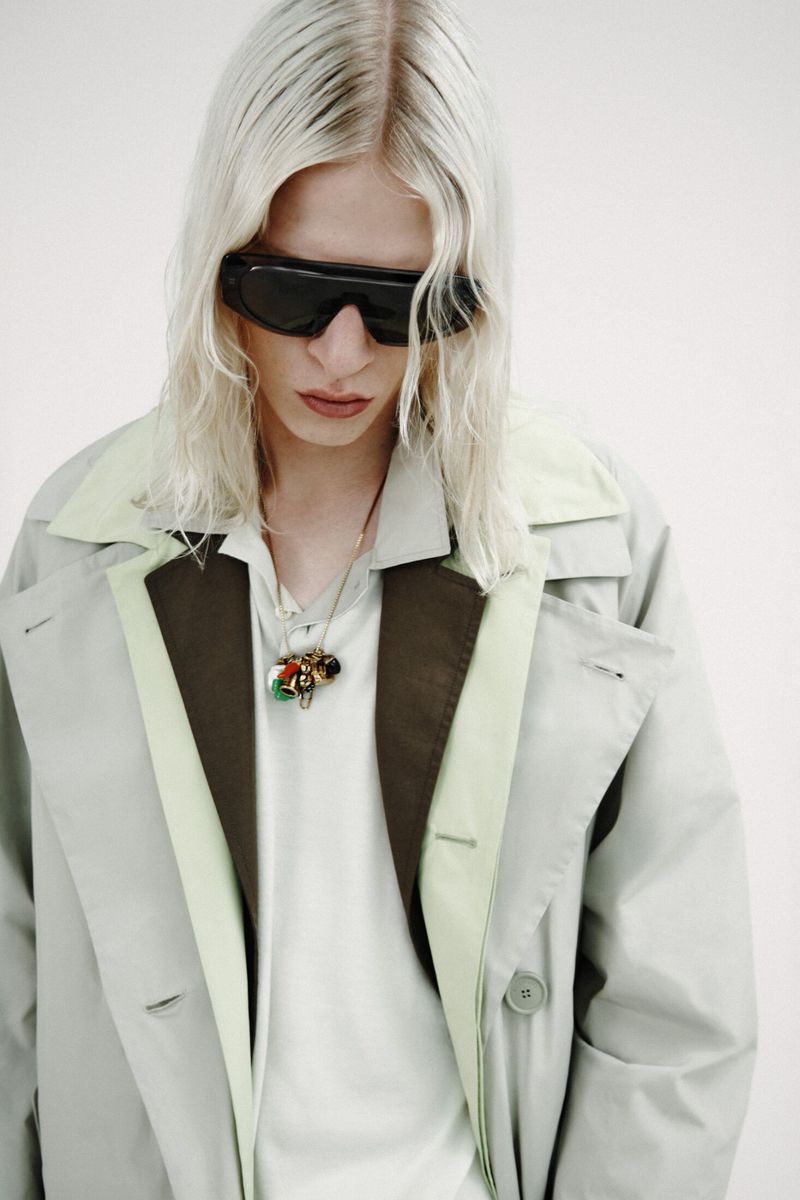Model with platinum blonde hair wearing dark wrap sunglasses and a layered light green coat against a white background.