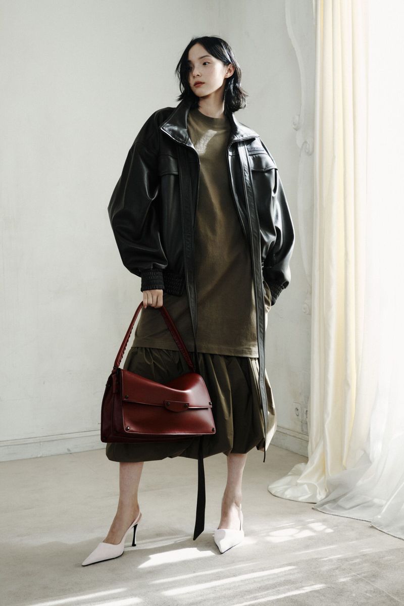 Model Xiao Wen Ju posing in an oversized black leather jacket, olive dress, and holding a burgundy leather handbag in a studio.
