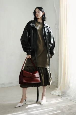 Model Xiao Wen Ju posing in an oversized black leather jacket, olive dress, and holding a burgundy leather handbag in a studio.