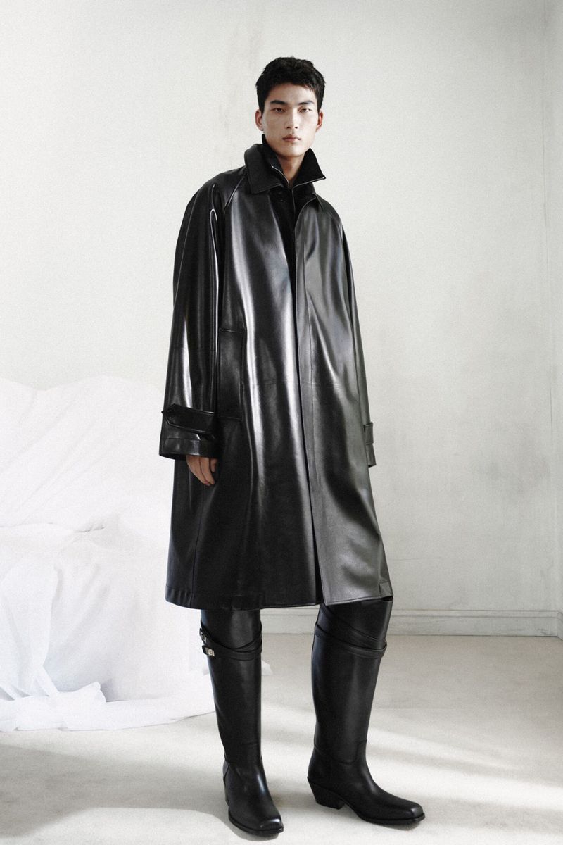 An East Asian male model stands wearing an oversized shiny black leather trench coat paired with tall black leather boots.