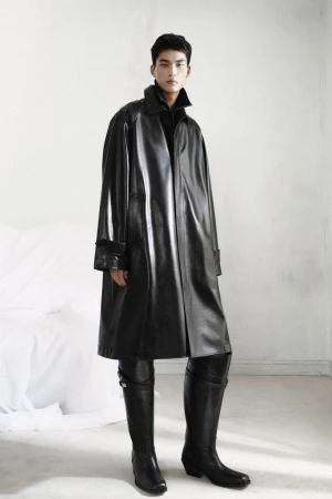 An East Asian male model stands wearing an oversized shiny black leather trench coat paired with tall black leather boots.