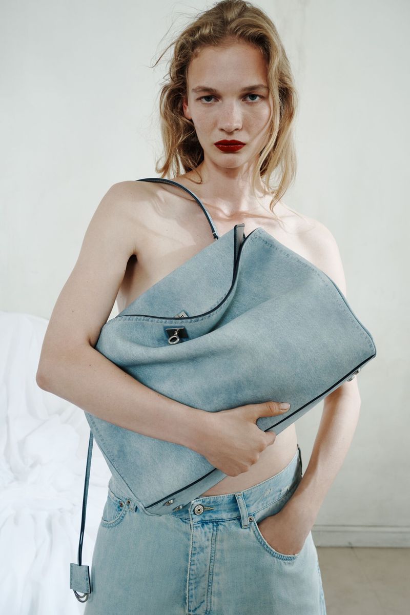 Model holding a large light blue denim tote bag against her torso while wearing matching denim jeans against a pale backdrop.