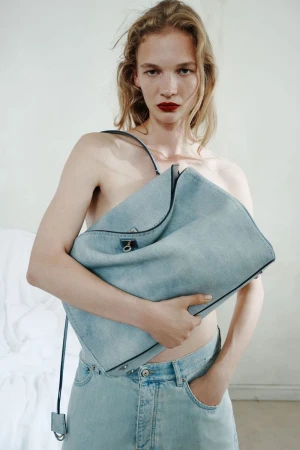 Model holding a large light blue denim tote bag against her torso while wearing matching denim jeans against a pale backdrop.